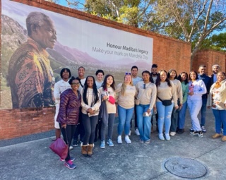 A group of students stand in front of a mural of Nelson Mandela against a mountainous landscape. The following text is written in black letters on the sky: Honour Madiba's legacy. Make your mark on Mandela Day.