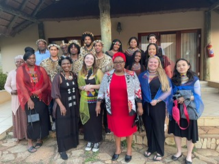 A group of people, some wearing traditional Zulu cultural attire, stand on a porch, all smiling. 